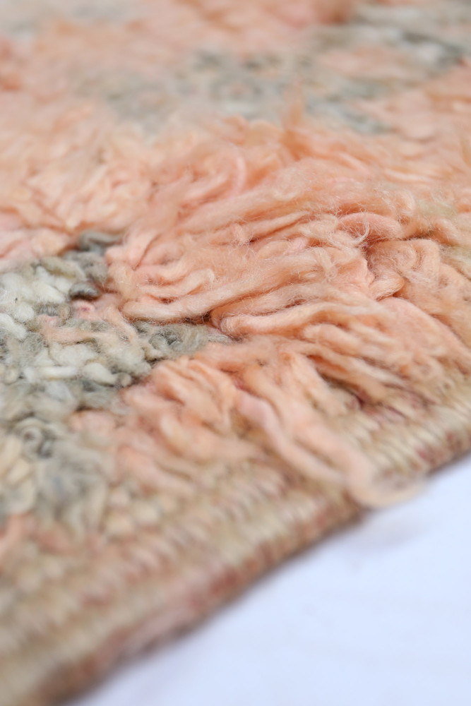 Tapis berbère aux teintes douces de rose et gris, avec des motifs tissés en laine composée de franges texturées.