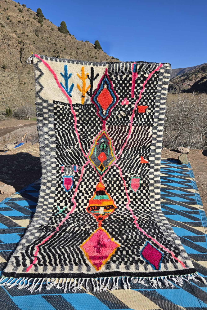 Tapis berbère au design géométrique coloré, avec des motifs vifs en rose, noir, et blanc sur un fond texturé.