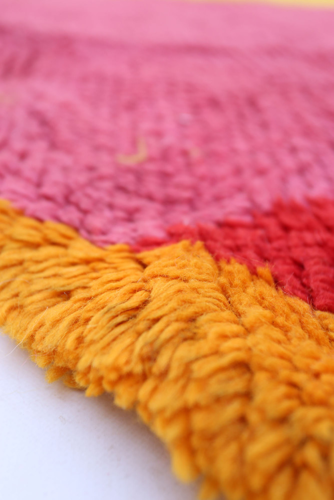 Tapis berbère coloré avec des motifs géométriques en rose, rouge et jaune, offrant une texture douce et chaleureuse.