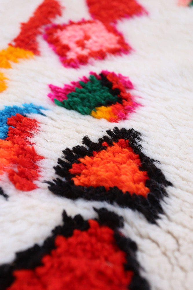 Tapis berbère aux motifs colorés avec des triangles noirs, des touches de rouge, rose et vert sur fond blanc.