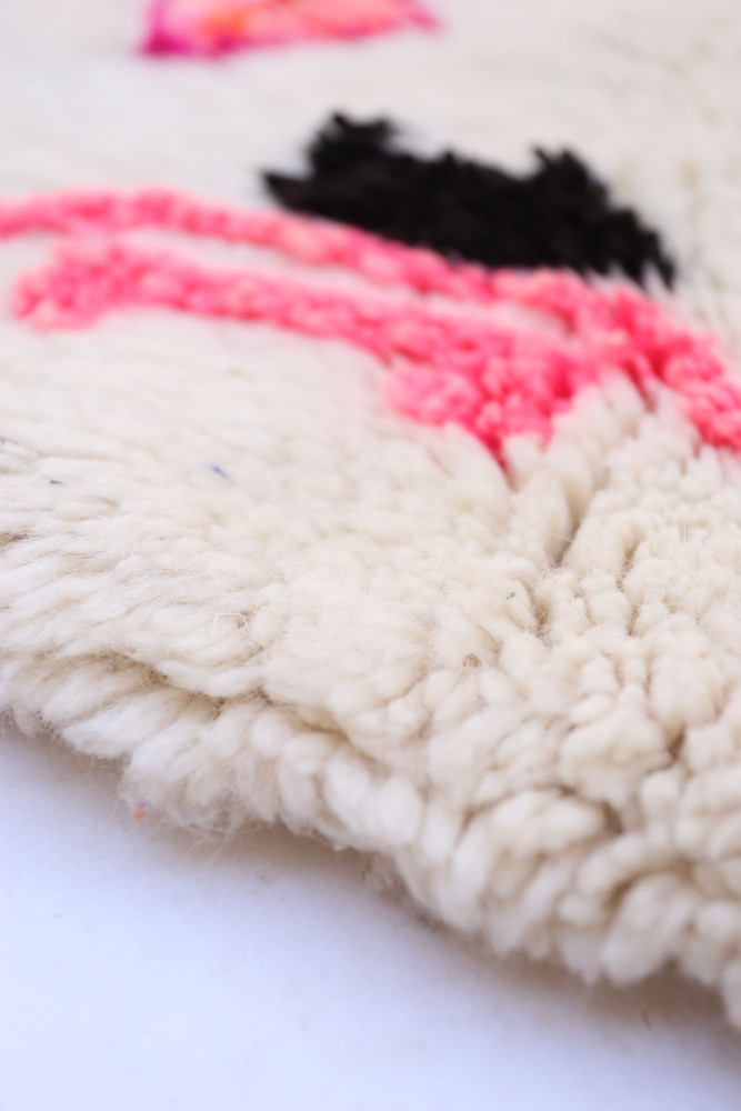 Tapis berbère en laine, couleurs crème avec motifs colorés en rose et noir, style artisanal unique.