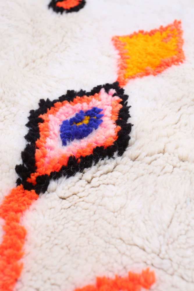 Tapis berbère au fond crémeux, avec des motifs colorés en rose, orange, bleu et noir, ajoutant une touche moderne et chaleureuse