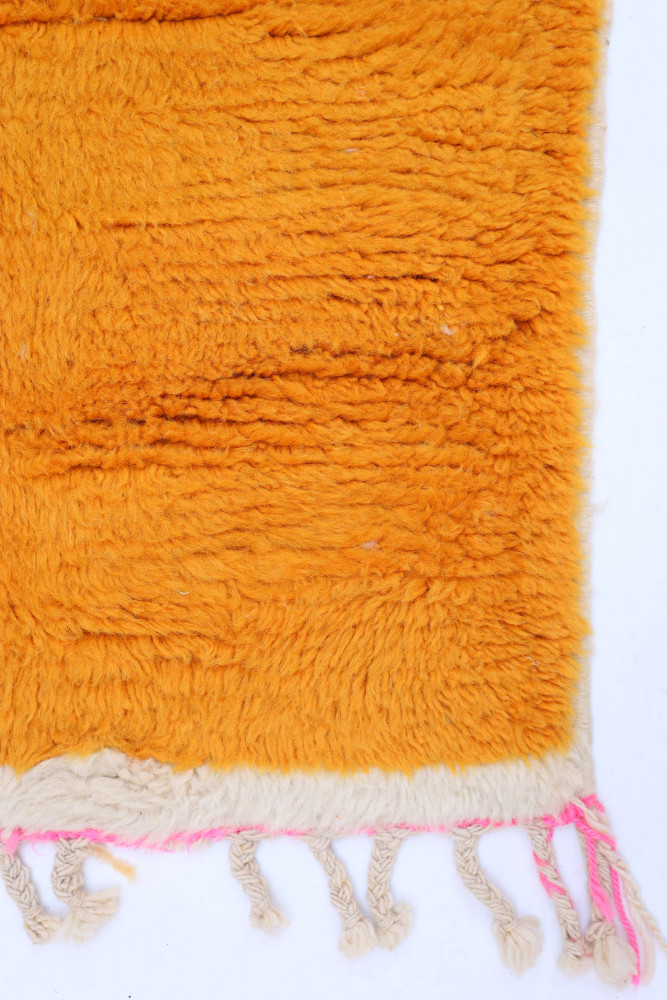 Tapis berbère orange vif avec des motifs en relief et des franges blanches et roses, ajoutant une touche artisanale.