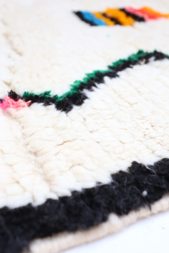 Tapis berbère beige avec des motifs géométriques colorés en noir, vert, rose et jaune, apportant une touche artisanale.