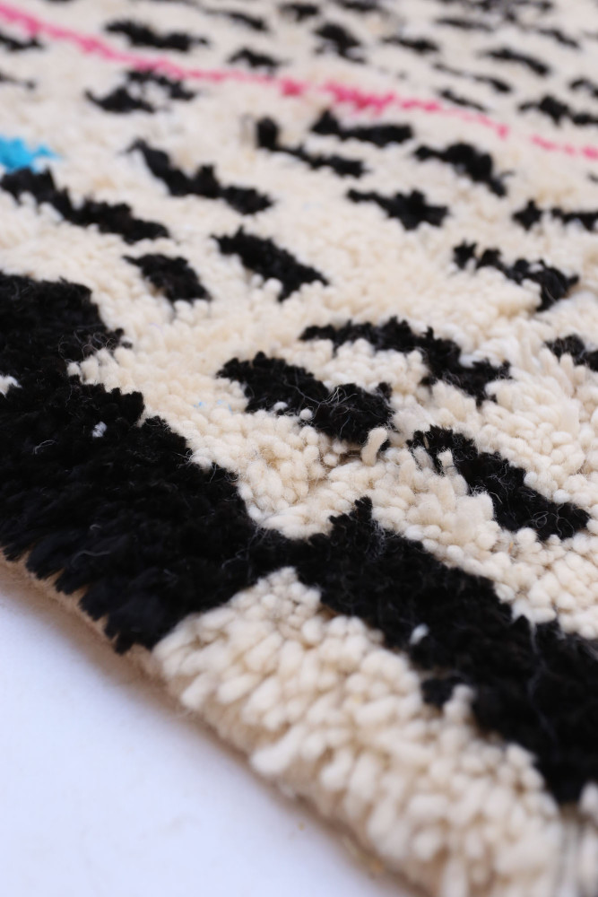 Tapis berbère doux avec motifs géométriques noirs et touches de rose et bleu sur fond crème.