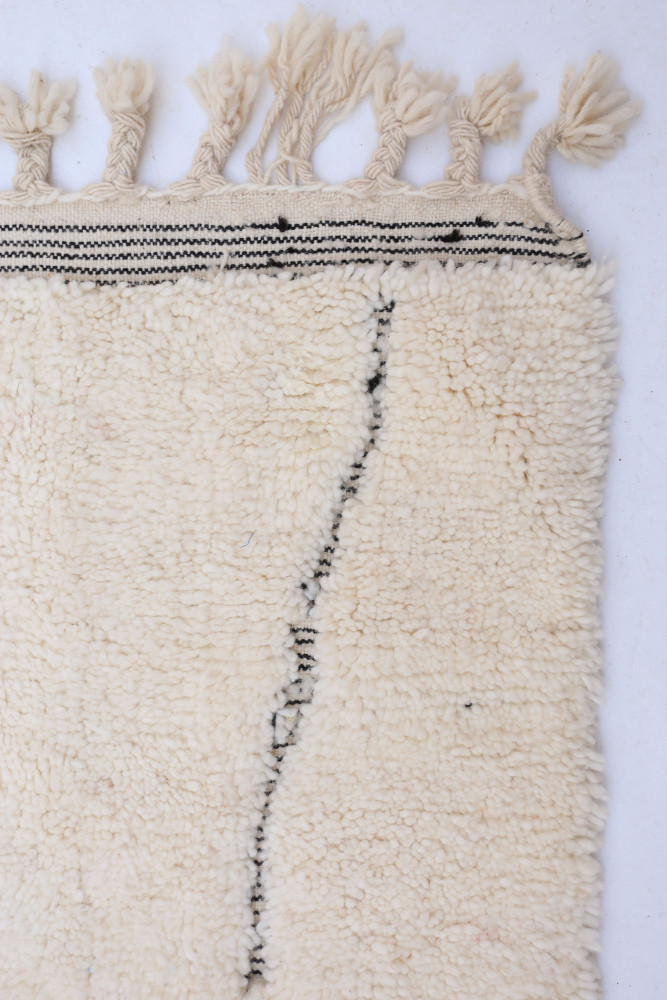 Tapis berbère en laine naturelle, couleur crème avec des motifs géométriques noirs et franges décoratives.