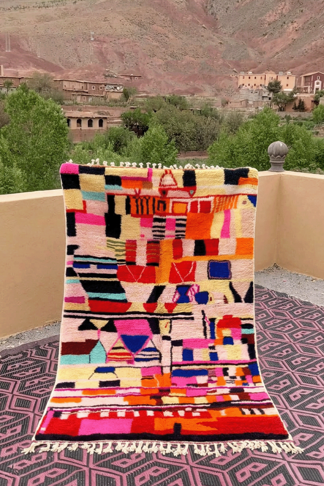 Tapis berbère coloré avec des motifs géométriques en rose, bleu, orange et noir sur fond clair.