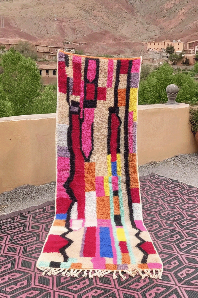 Tapis berbère coloré avec des motifs géométriques en rose, jaune, violet et noir sur fond crème.