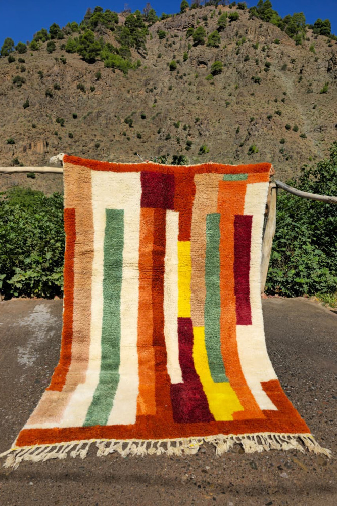 Tapis berbère aux motifs géométriques colorés, dominé par des teintes terracotta, vertes et jaunes sur fond beige.