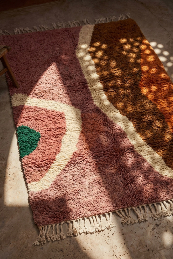 Tapis berbère aux teintes roses, marron et vert, avec des motifs géométriques modernes et des franges traditionnelles.
