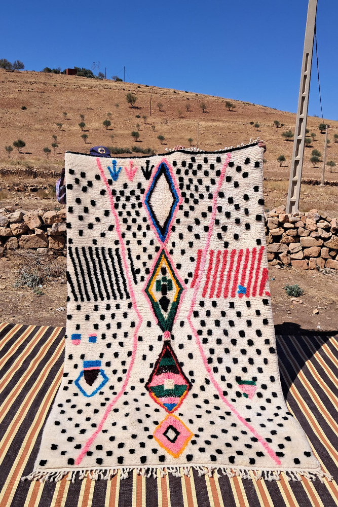 Tapis berbère aux motifs géométriques colorés sur fond crème, avec des touches de rose, noir et vert.