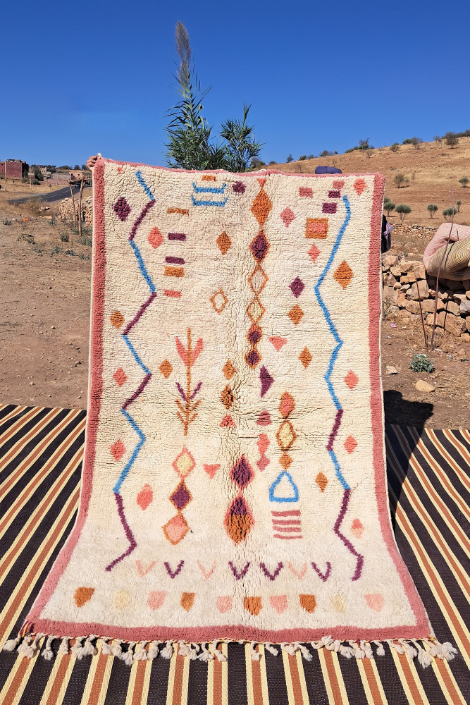 Tapis berbère en laine, style traditionnel avec des motifs géométriques en bleu, orange et violet sur fond crème.