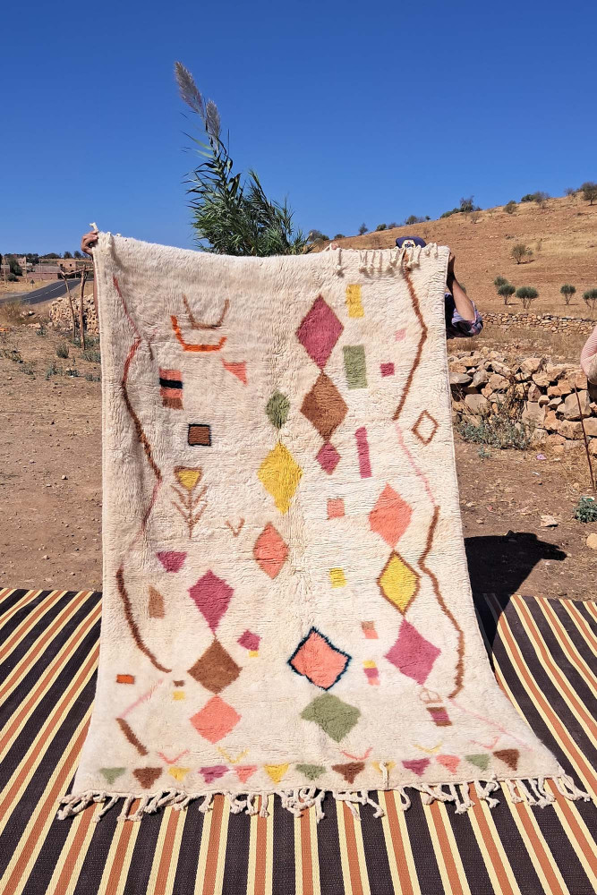 Tapis berbère aux motifs géométriques colorés en rose, jaune et vert sur fond crème, parfait pour une décoration chaleureuse.