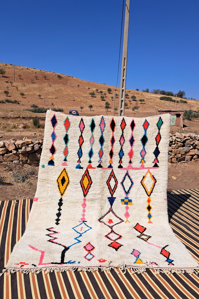 Tapis berbère blanc avec motifs colorés en losanges et lignes géométriques dans les teintes roses, jaunes et bleues.
