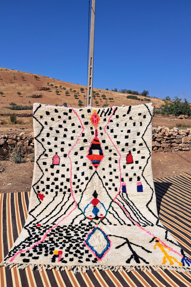 Tapis berbère en laine, couleurs vives, motifs géométriques noirs et blancs avec des touches de rose et bleu.