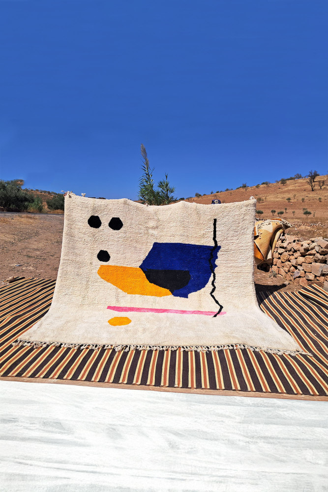 Tapis berbère coloré avec des motifs géométriques en bleu, noir, jaune et rose sur fond crème.