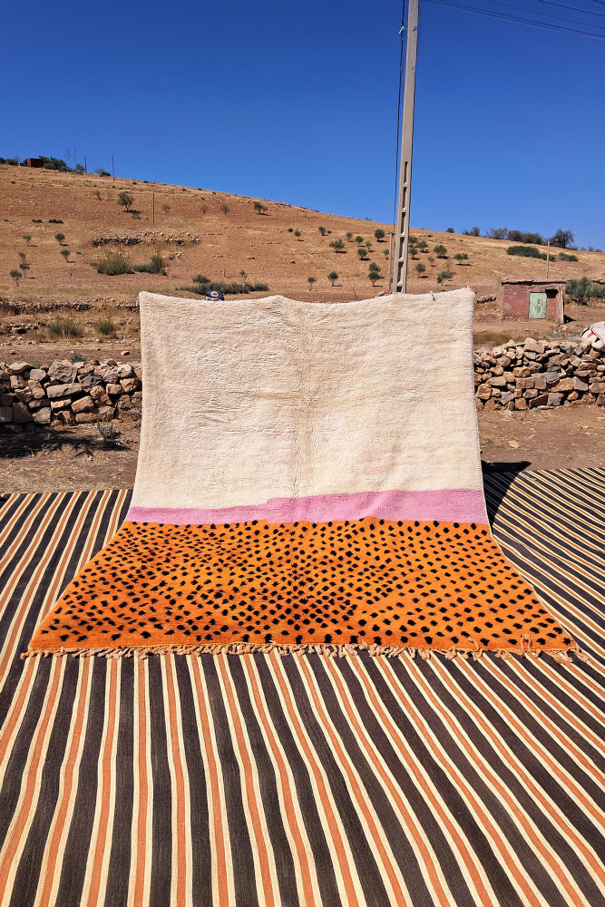 Tapis berbère en laine beige avec des motifs géométriques orange et roses sur un fond texturé.