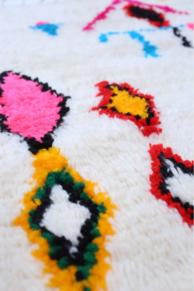 Tapis berbère coloré avec des motifs géométriques en rose, noir, jaune et bleu sur un fond blanc doux.
