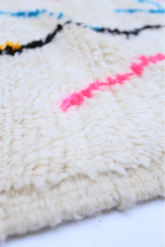Tapis berbère en laine, couleur crème avec des motifs géométriques en bleu, rose, et noir.