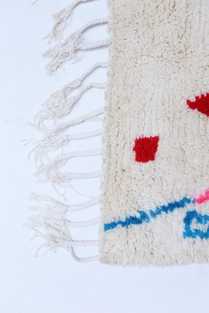 Tapis berbère aux motifs colorés en rouge et bleu sur un fond beige, avec des franges décoratives au bord.