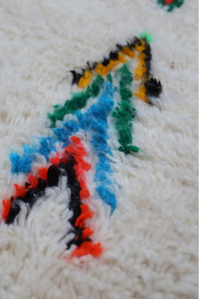 Tapis berbère aux motifs géométriques colorés avec des teintes blanc, bleu, rouge et noir sur un fond crémeux.