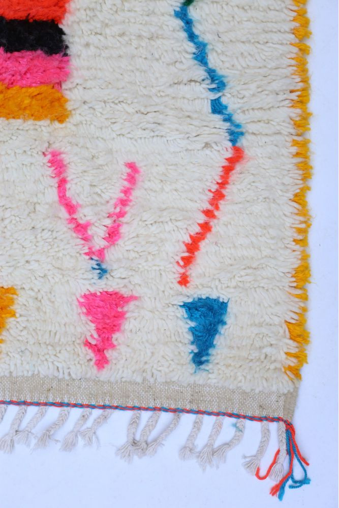 Tapis berbère à motifs géométriques colorés, dominé par le blanc avec des touches de rose, bleu et orange.